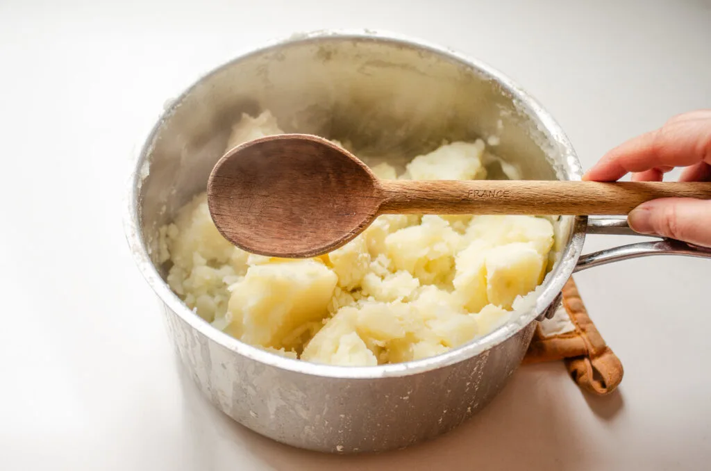 A hand holding a wooden spoon over a pot of boiled potatoes.