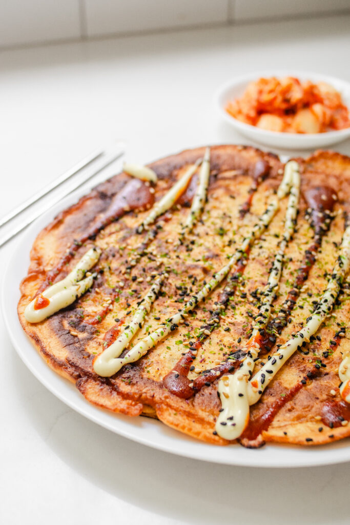 A sourdough discard kimchi pancake on a dinner plate with a bowl of kimchi in the background.