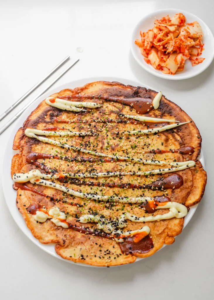 A sourdough discard kimchi pancake on a dinner plate with a bowl of kimchi in the background.