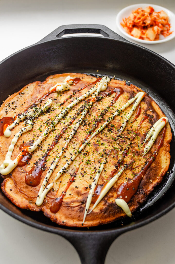 A sourdough discard kimchi pancake in a cast iron skillet with mayonnaise, sriracha, and furikake on top with a bowl of kimchi off to the side.