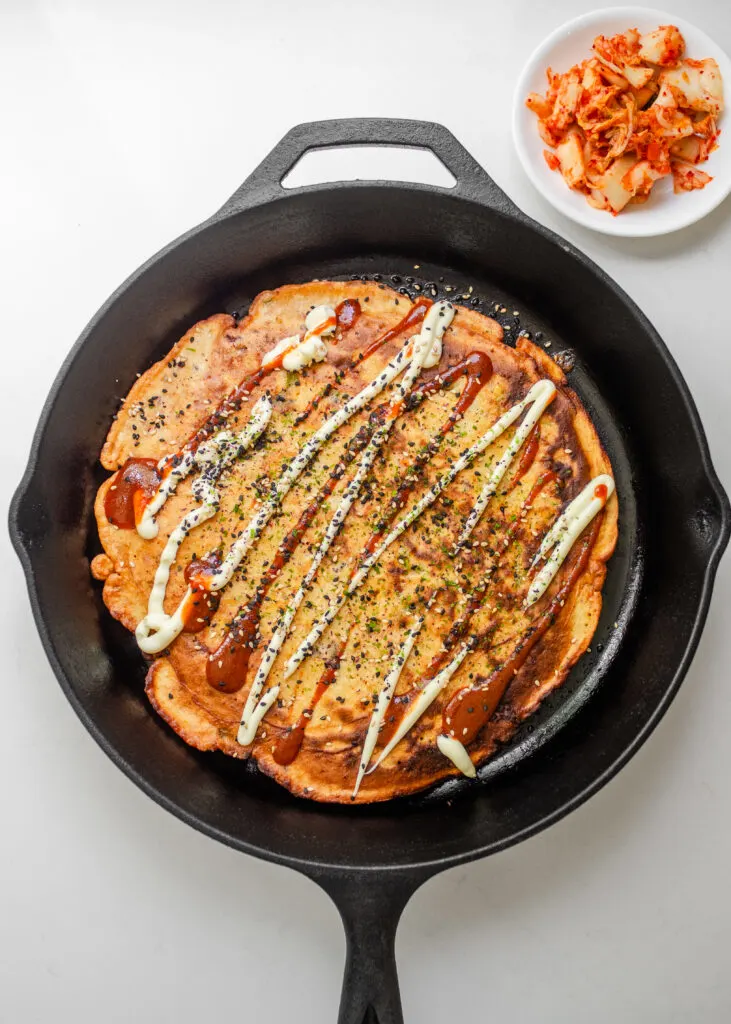 A sourdough discard kimchi pancake in a cast iron skillet with mayonnaise, sriracha, and furikake on top with a bowl of kimchi off to the side.