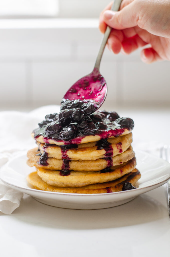 Scooping blueberry compote made with fresh blueberries on top of a stack of pancakes.