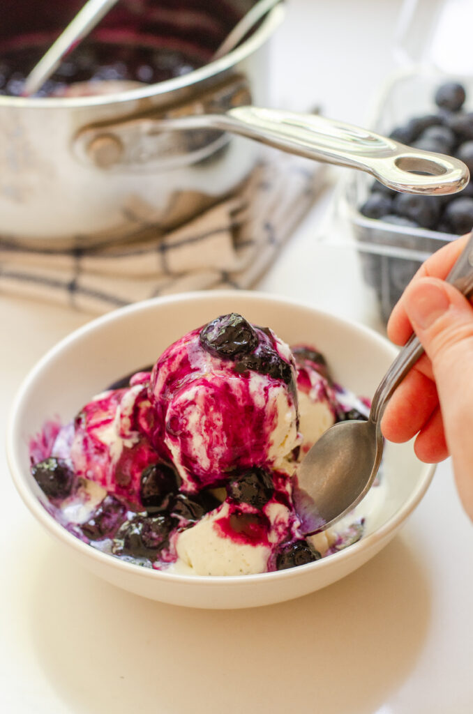Warm blueberry compote on top of a bowl of vanilla ice cream with the saucepan and fresh blueberries behind it.