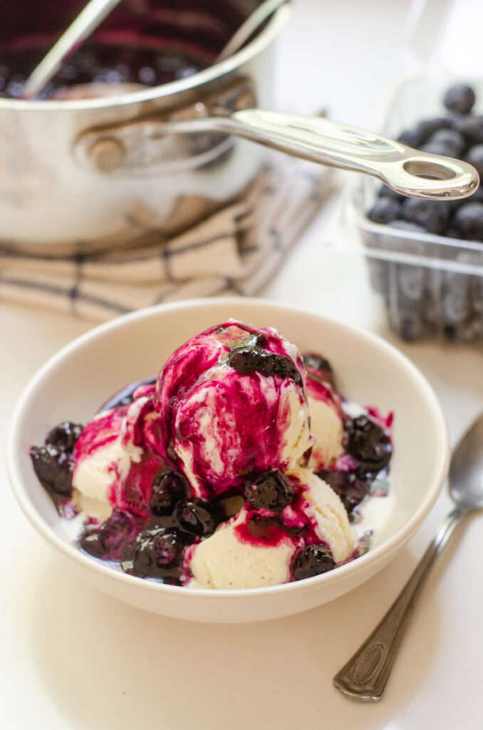 Warm blueberry compote on top of a bowl of vanilla ice cream with the saucepan and fresh blueberries behind it.