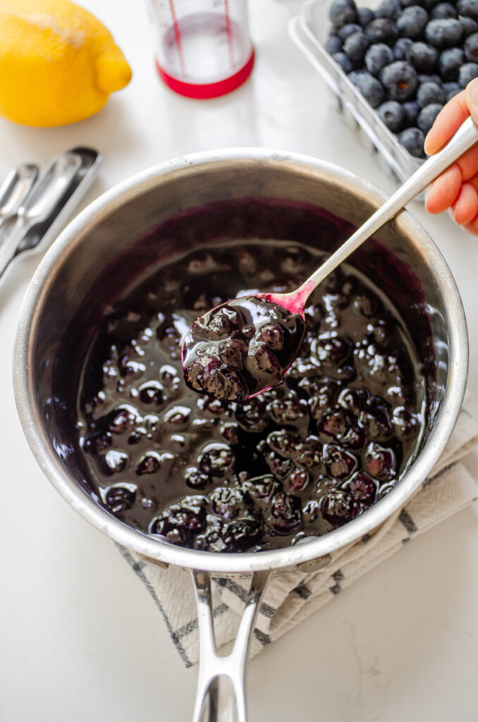 A pan of blueberry compote with a hand holding a spoonful with fresh blueberries behind the pan.