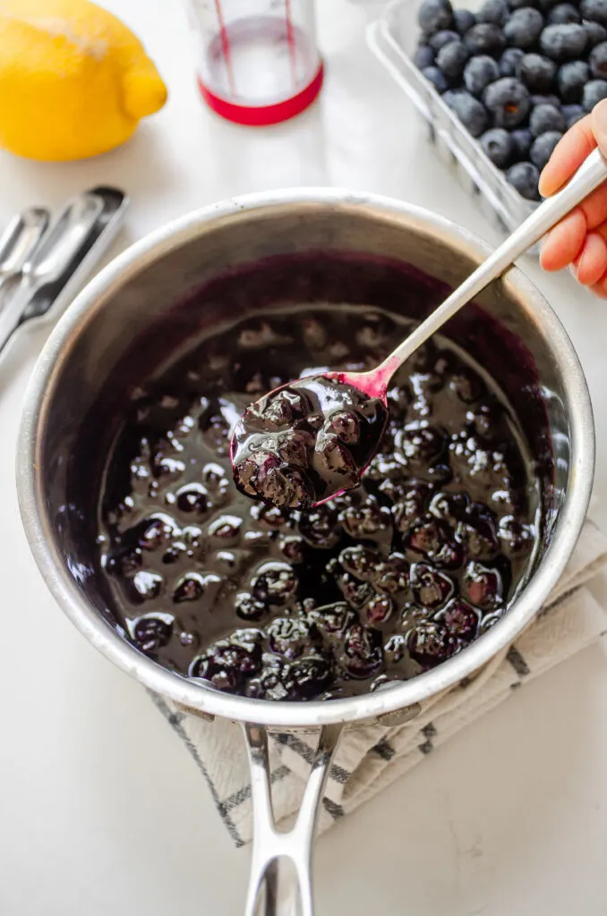 A pan of blueberry compote with a hand holding a spoonful with fresh blueberries behind the pan.