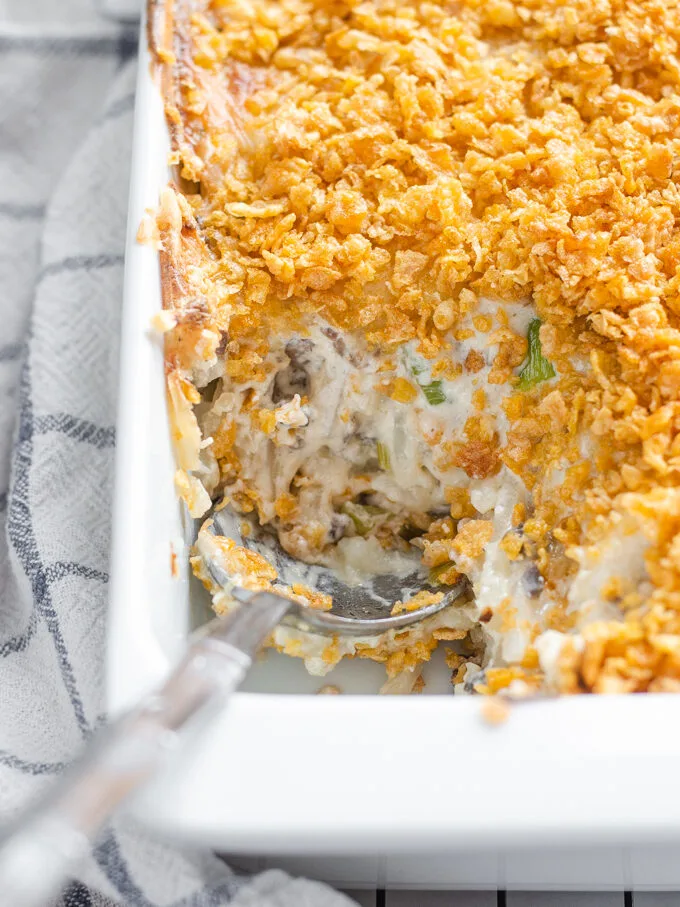 The baked gluten free cheesy potatoes (AKA funeral potatoes) in a white casserole dish on top of a wire cooling rack with a scoop taken out.