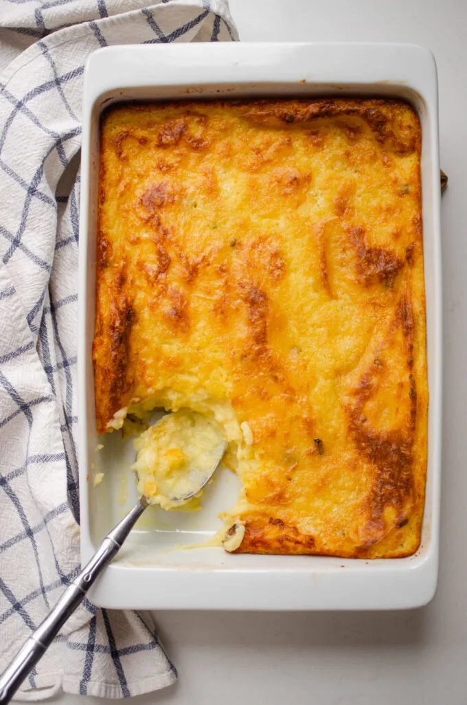 A pan of baked mashed potatoes with a scoop taken out and a serving spoon in the pan with a tea towel off to the side.