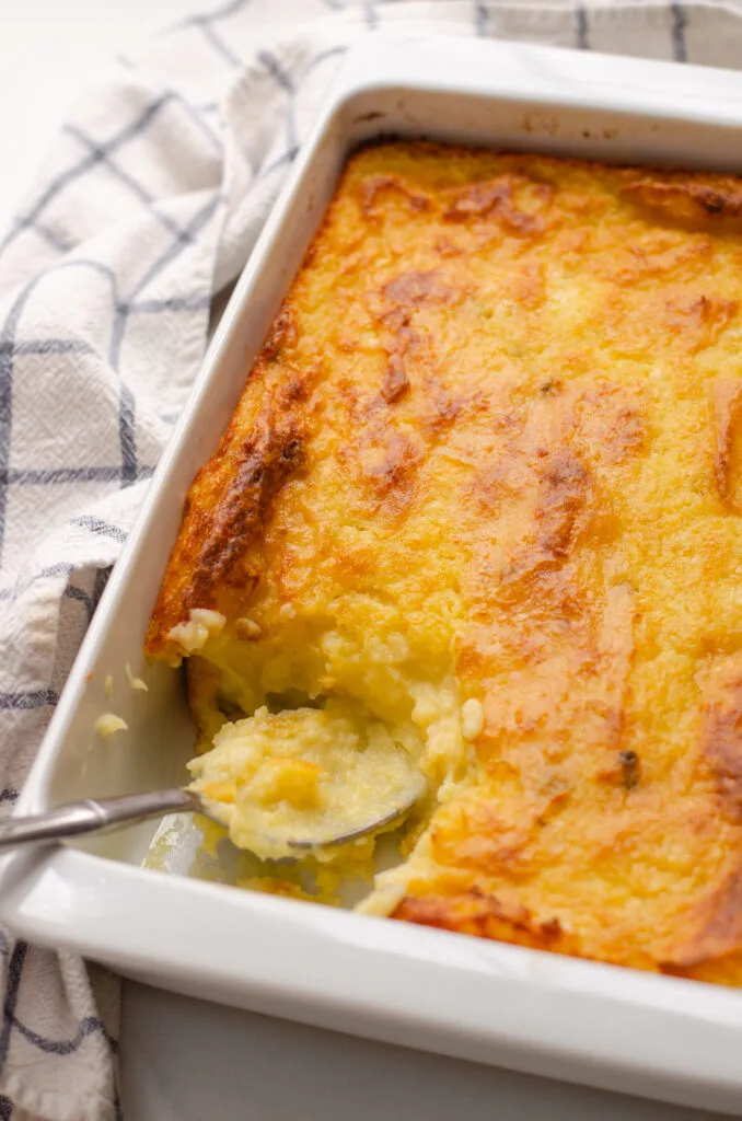 A pan of baked mashed potatoes with a scoop taken out and a serving spoon in the pan with a tea towel off to the side.