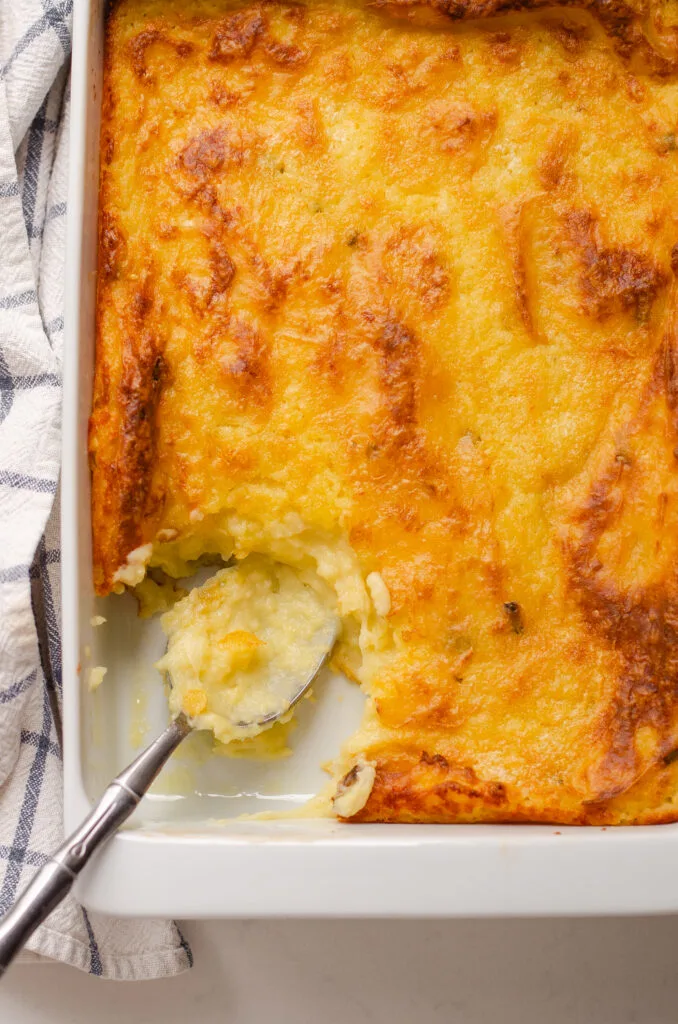 A pan of baked mashed potatoes with a scoop taken out and a serving spoon in the pan with a tea towel off to the side.