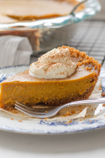 A slice of pumpkin pie with a graham cracker crust on a vintage blue and white plate with the whole pie in the background.