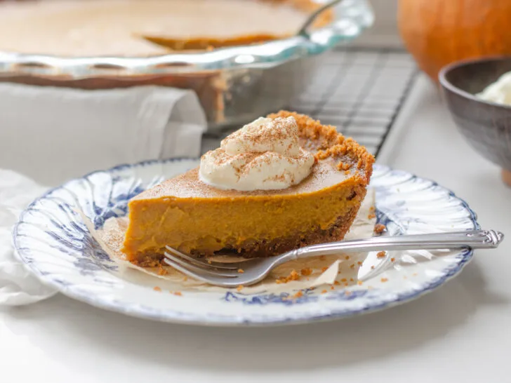 A slice of pumpkin pie with a graham cracker crust on a vintage blue and white plate with the whole pie in the background.
