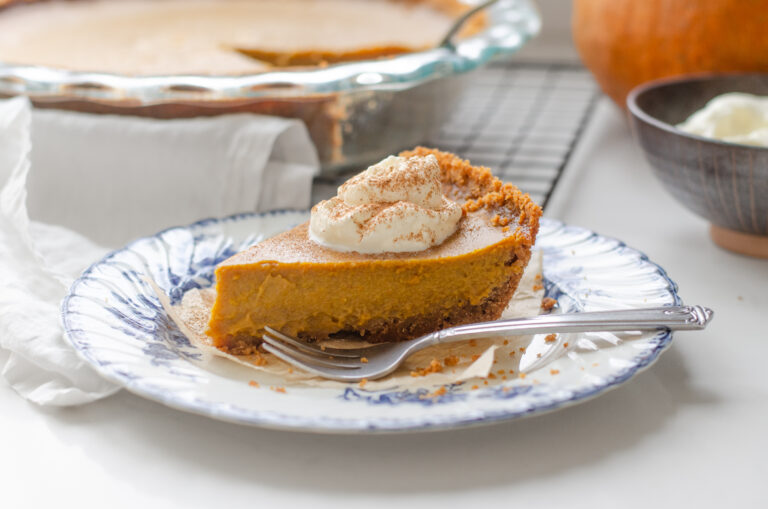 A slice of pumpkin pie with a graham cracker crust on a vintage blue and white plate with the whole pie in the background.