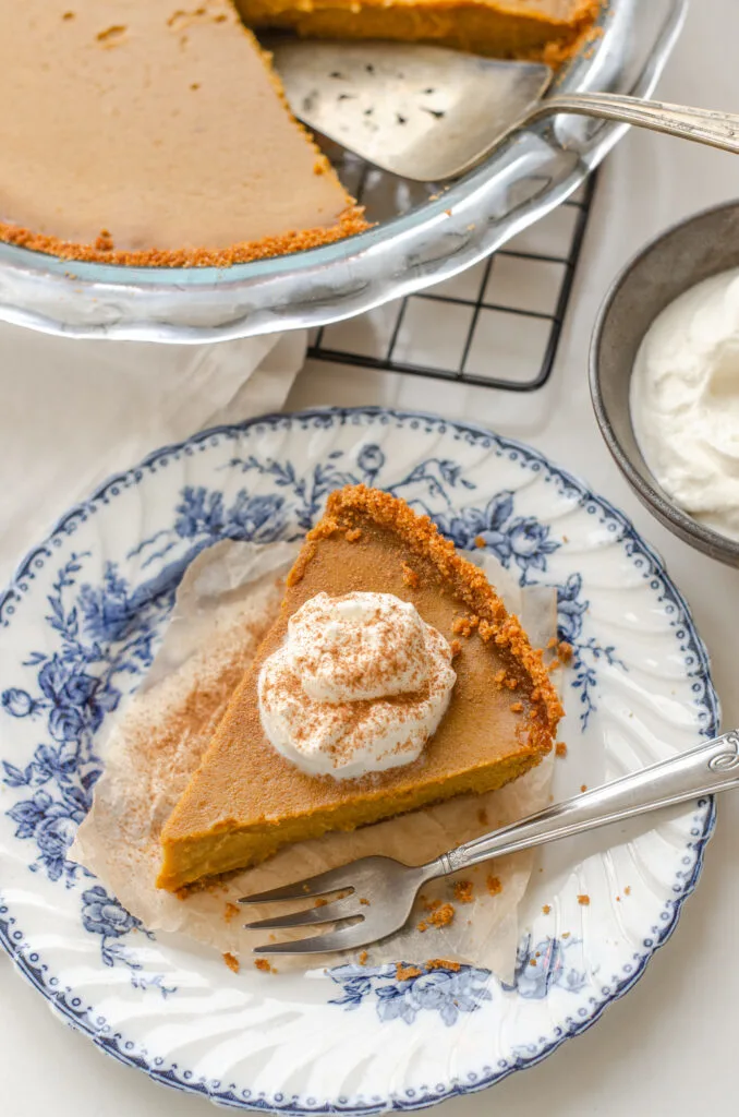 A slice of pumpkin pie with a graham cracker crust on a vintage blue and white plate with the whole pie in the background.