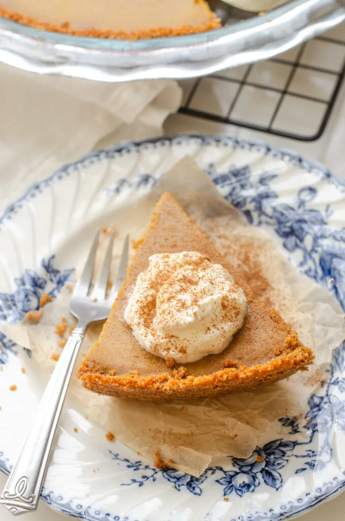 A slice of pumpkin pie with a graham cracker crust on a vintage blue and white plate with the whole pie in the background.