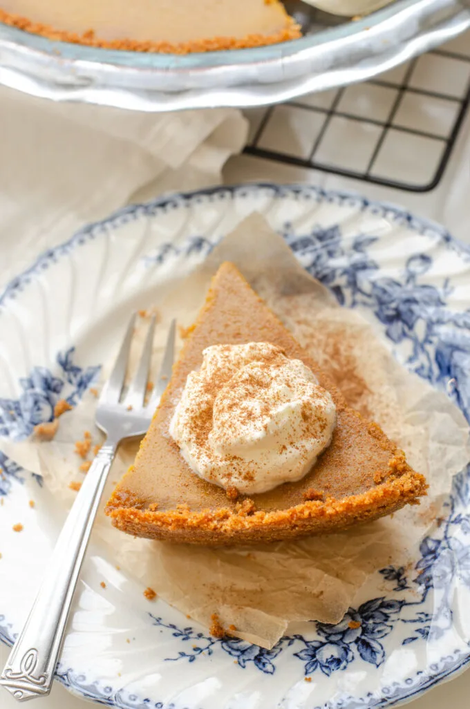 A slice of pumpkin pie with a graham cracker crust on a vintage blue and white plate with the whole pie in the background.