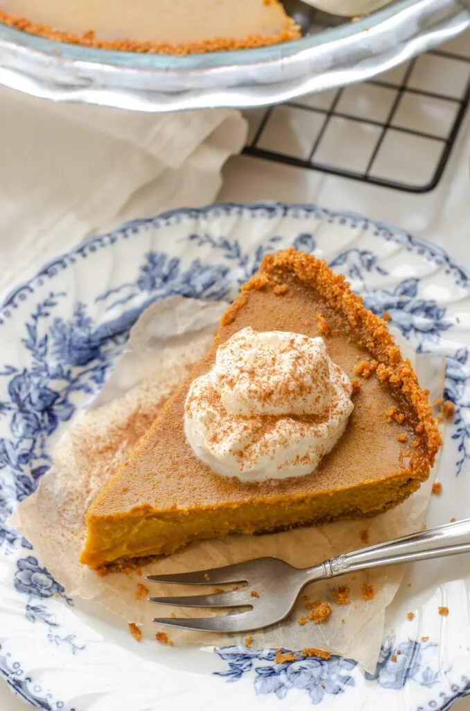 A slice of pumpkin pie with a graham cracker crust on a vintage blue and white plate with the whole pie in the background.