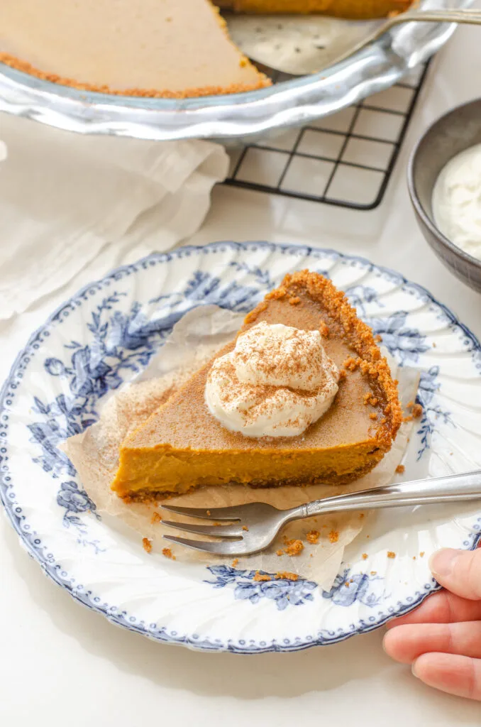 A piece of pumpkin pie with a graham cracker crust on a blue and white vintage plate with a dollop of whipped cream on top.