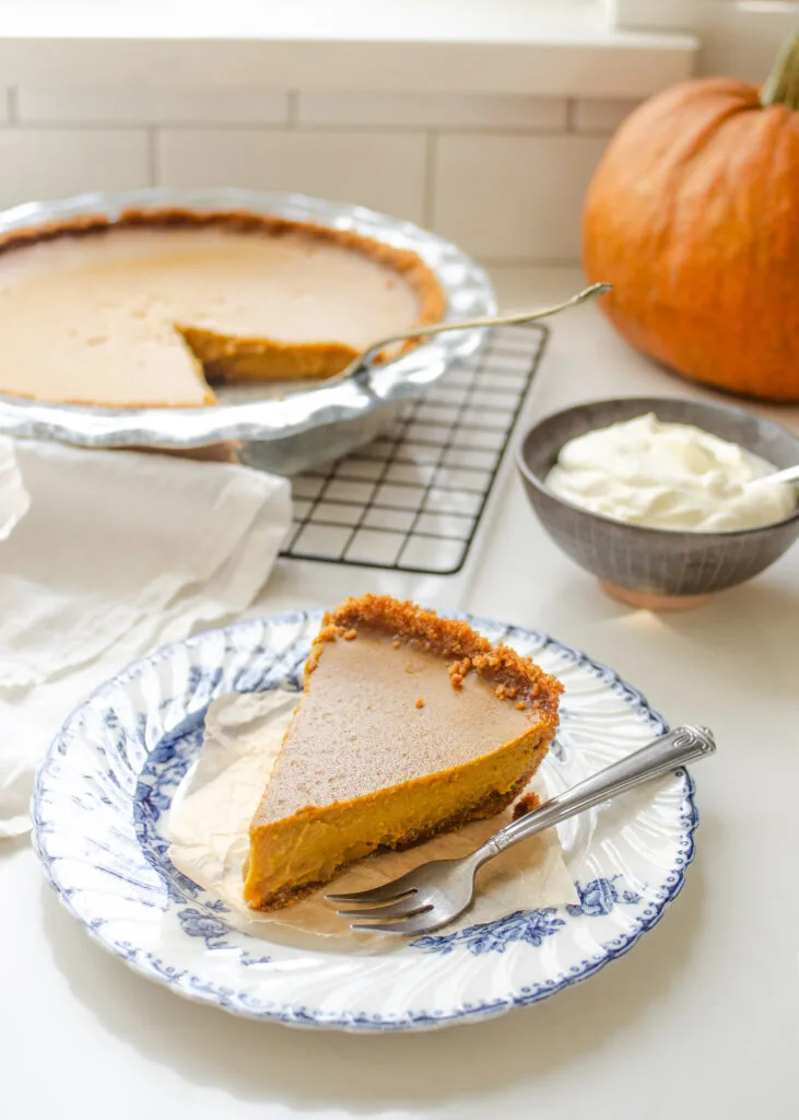A slice of pumpkin pie with a graham cracker crust on a vintage blue and white plate with the whole pie in the background.
