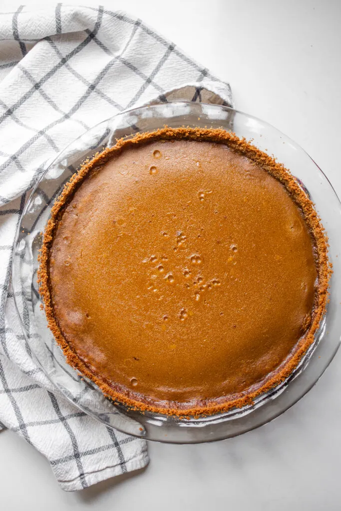The baked pumpkin pie on a white countertop with a checked dish towel to the side.