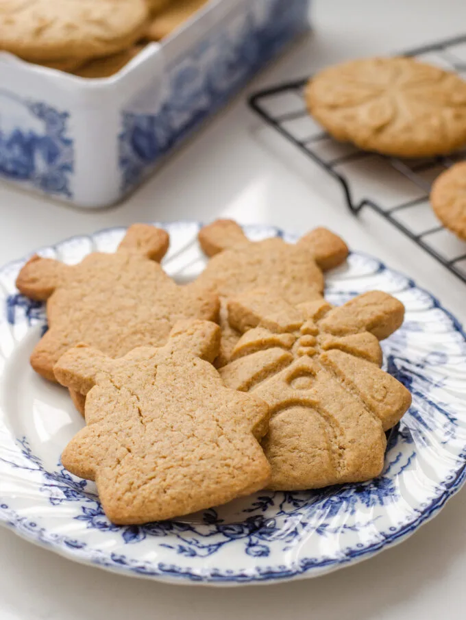 Speculoos cookies on a blue and white plate with more cookies in a tin behind them, and on a wire rack to the side.