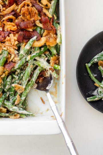 A pan of green bean casserole with bacon and fried onions with a scoop taken out and the plate with the scoop next to the pan.