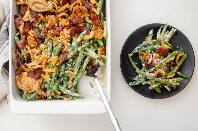 A pan of green bean casserole with bacon and fried onions with a scoop taken out and the plate with the scoop next to the pan.