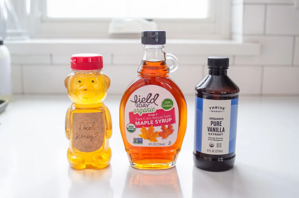 A jar of honey and maple syrup on a white countertop with a bottle of vanilla extract next to them.