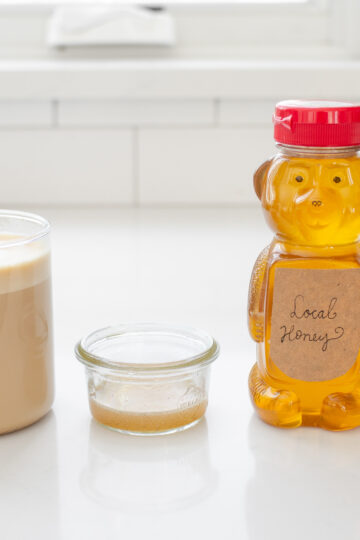 A latte, homemade vanilla syrup substitute, honey, and vanilla extract on a white countertop.