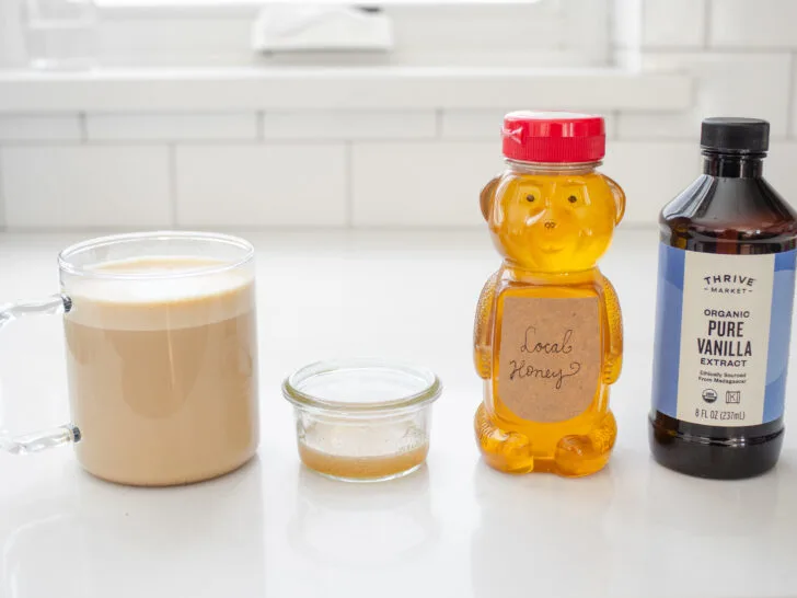 A latte, homemade vanilla syrup substitute, honey, and vanilla extract on a white countertop.