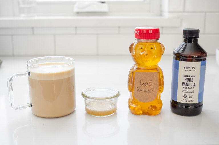 A latte, homemade vanilla syrup substitute, honey, and vanilla extract on a white countertop.