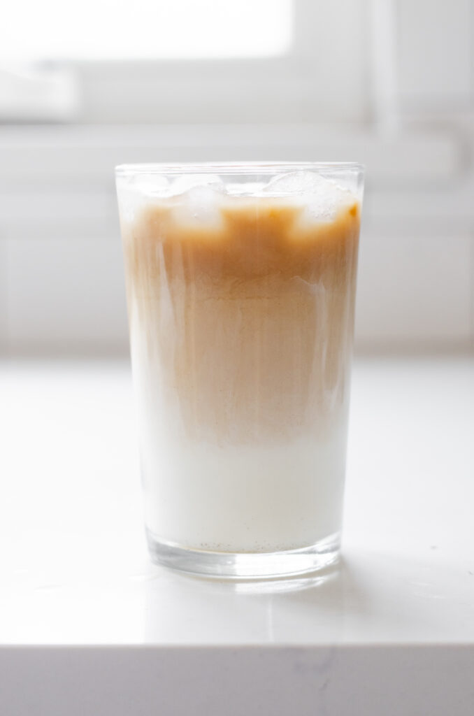 An iced London Fog in a transparent glass on a white countertop with a window in the background.