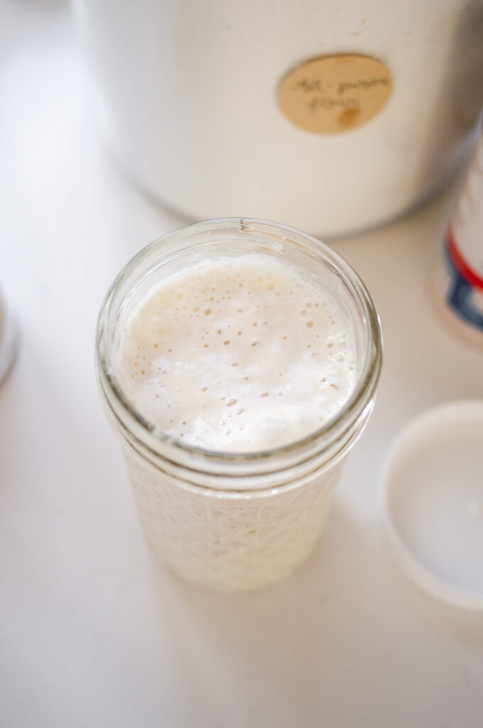 A jar of sourdough starter on a white countertop with a jar of flour behind it.
