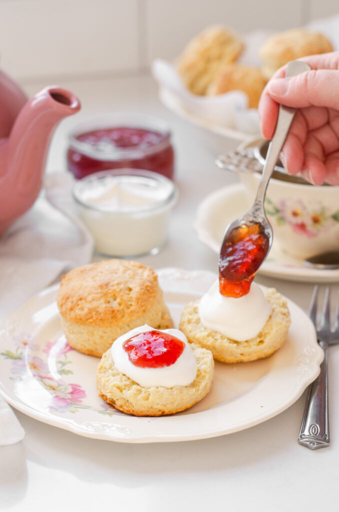 Sourdough scones on a fancy plate with a hand spoon jam on top of one of the scones, and a teapot and teacup in the background.