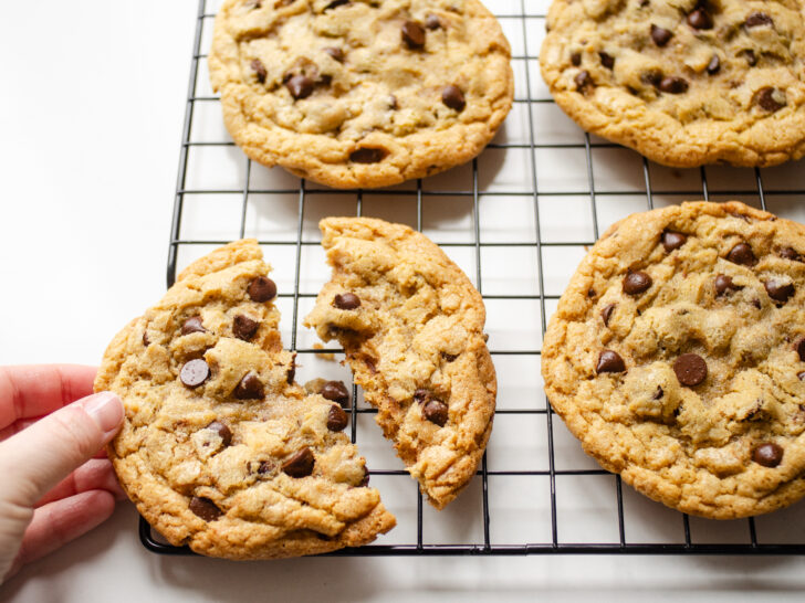 A hand holding a broken einkorn flour chocolate chip cookie on a wire cooling rack.