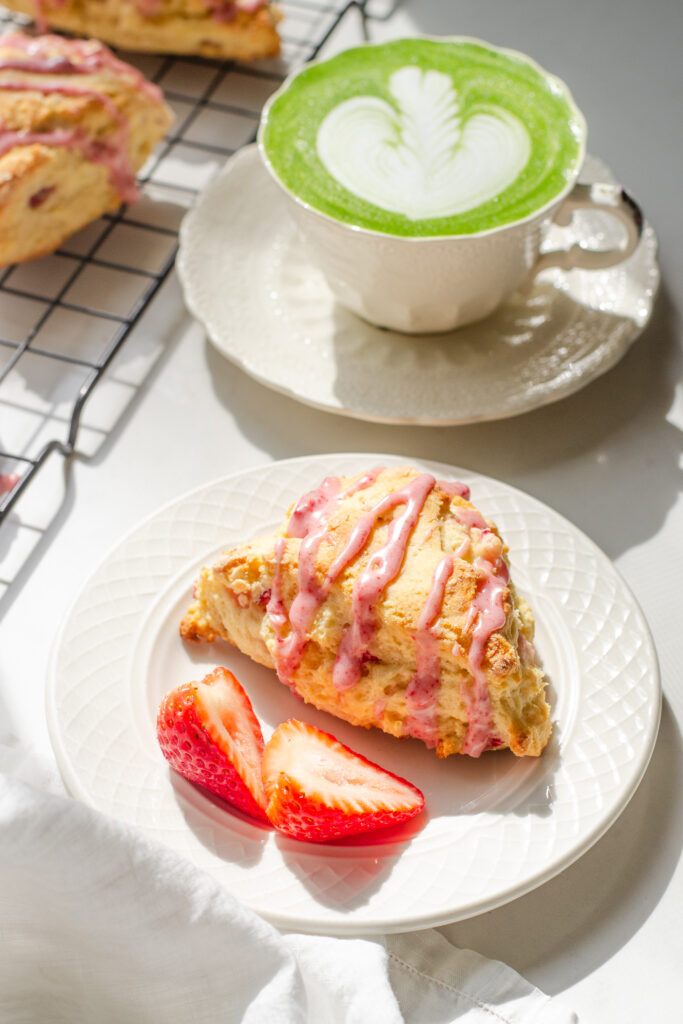 A strawberry scone on a plate with fresh strawberries beside it and a cup with tea in the background.