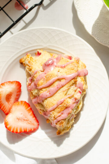 A strawberry scone on a plate with fresh strawberries beside it and a cup with tea in the background.