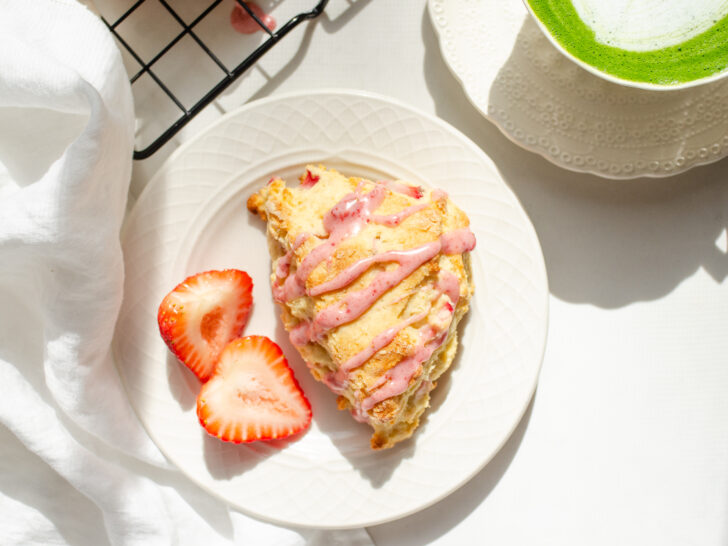 A strawberry scone on a plate with fresh strawberries beside it and a cup with tea in the background.