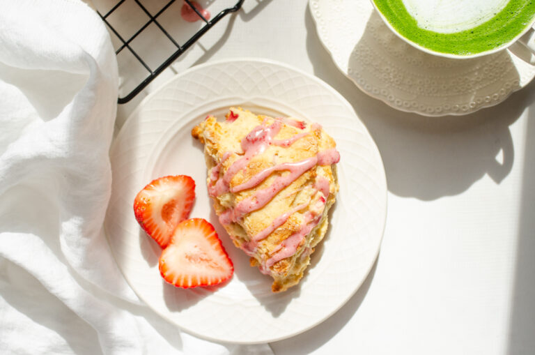 A strawberry scone on a plate with fresh strawberries beside it and a cup with tea in the background.