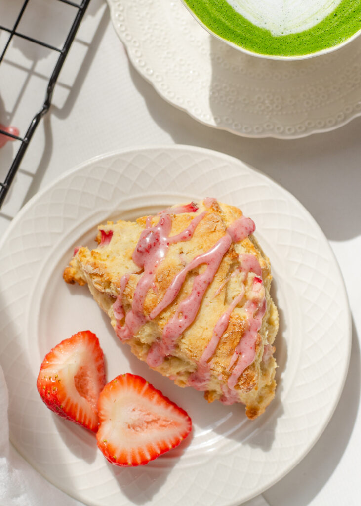 A strawberry scone on a plate with fresh strawberries beside it and a cup with tea in the background.