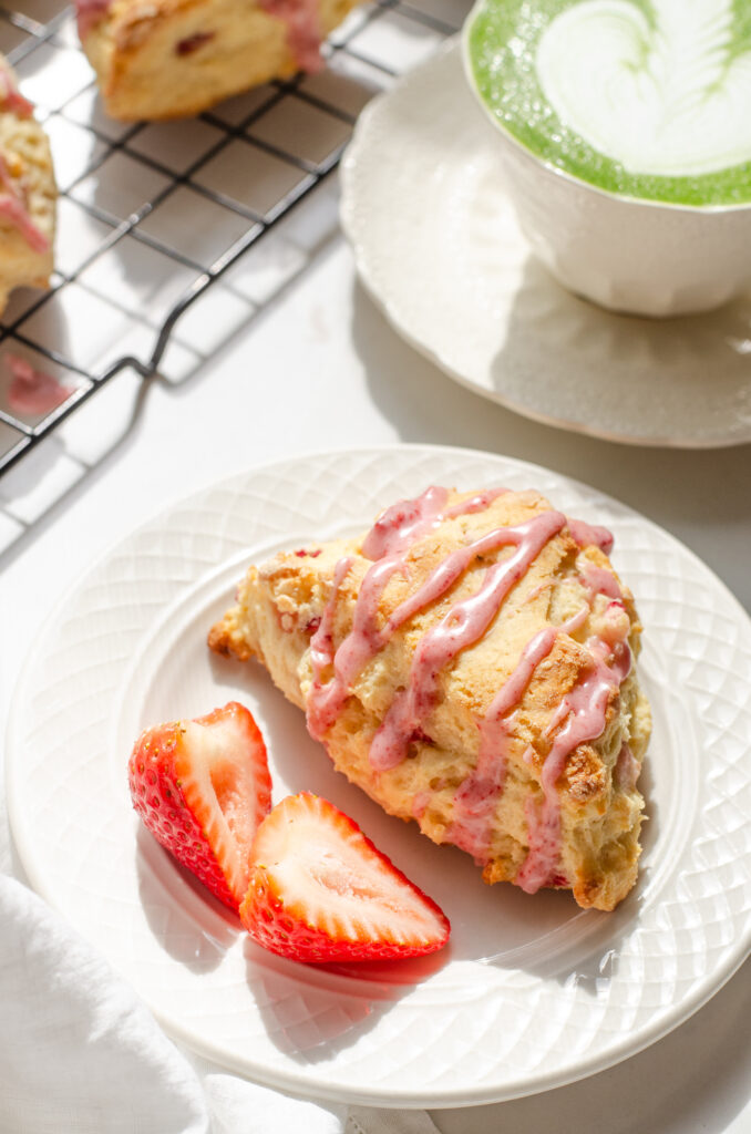 A strawberry scone on a plate with fresh strawberries beside it and a cup with tea in the background.