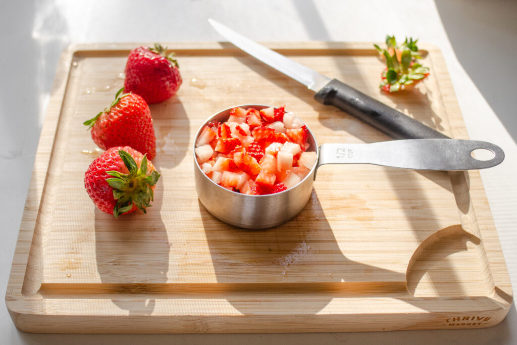 A measuring cup full of chopped strawberries on a wooden cutting board with more fresh strawberries and a knife in the background.