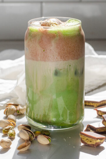 A copycat Starbucks Iced Dubai Chocolate Matcha latte on a sunny white countertop with pistachios on one side, and Dubai chocolate bars on the other.