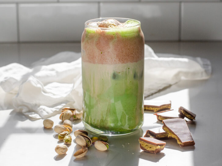 A copycat Starbucks Iced Dubai Chocolate Matcha latte on a sunny white countertop with pistachios on one side, and Dubai chocolate bars on the other.