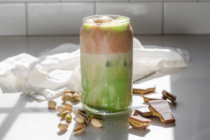 A copycat Starbucks Iced Dubai Chocolate Matcha latte on a sunny white countertop with pistachios on one side, and Dubai chocolate bars on the other.