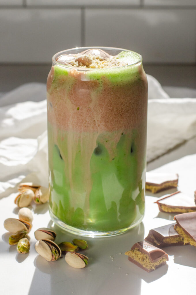 A copycat Starbucks Iced Dubai Chocolate Matcha latte on a sunny white countertop with pistachios on one side, and Dubai chocolate bars on the other.