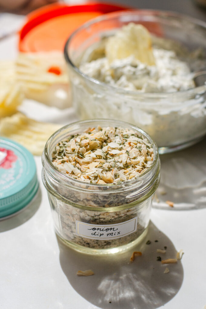 Onion dip mix in a jar on a sunny countertop with a bowl of the dip in the background.