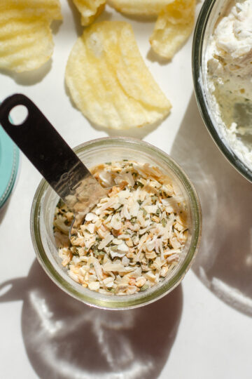 A jar of homemade onion dip mix on a sunny countertop with a container of the dip off to the side with a few potato chips around it.