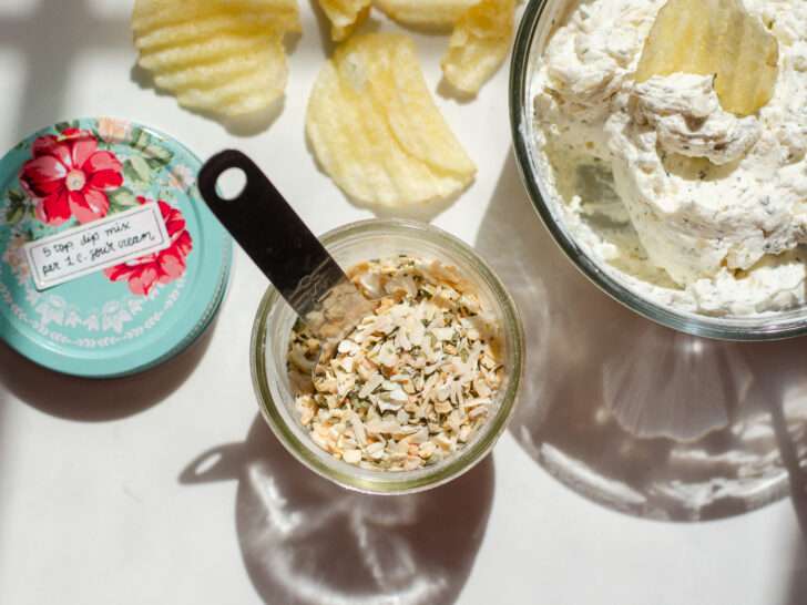 A jar of homemade onion dip mix on a sunny countertop with a container of the dip off to the side with a few potato chips around it.