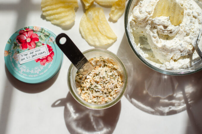 A jar of homemade onion dip mix on a sunny countertop with a container of the dip off to the side with a few potato chips around it.